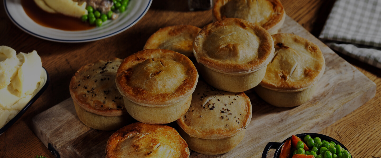 A selection of Brewers Fayre pies stacked on a wooden board with a bowl of mashed potatoes and peas.