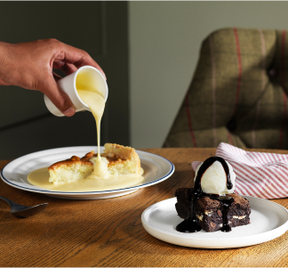 Custard being poured onto a slice of apple crumble pie and a chocolate brownie with vanilla ice cream on a table.