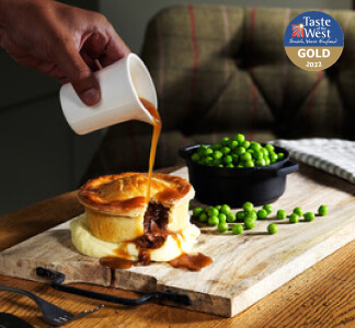A jug of gravy being poured onto the Brewers Fayre Beef and Ale pie on mashed potato and a dish of green peas.